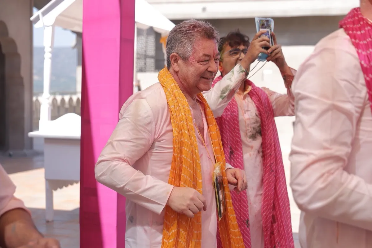 A colour-dusted guest grinning in an orange dupatta while a friend films