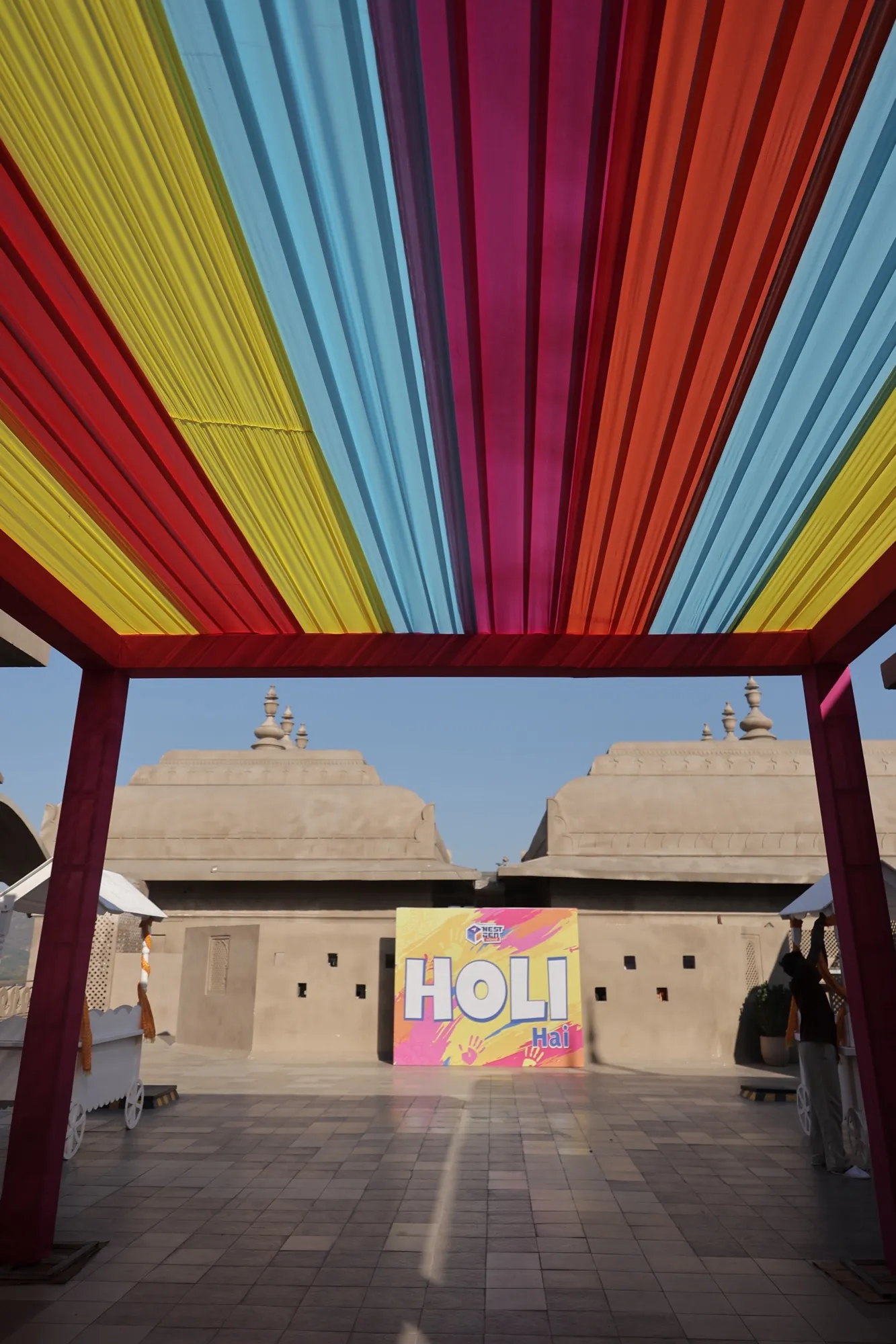 A rainbow canopy of coloured cloth over the empty rooftop, the Holi sign beyond