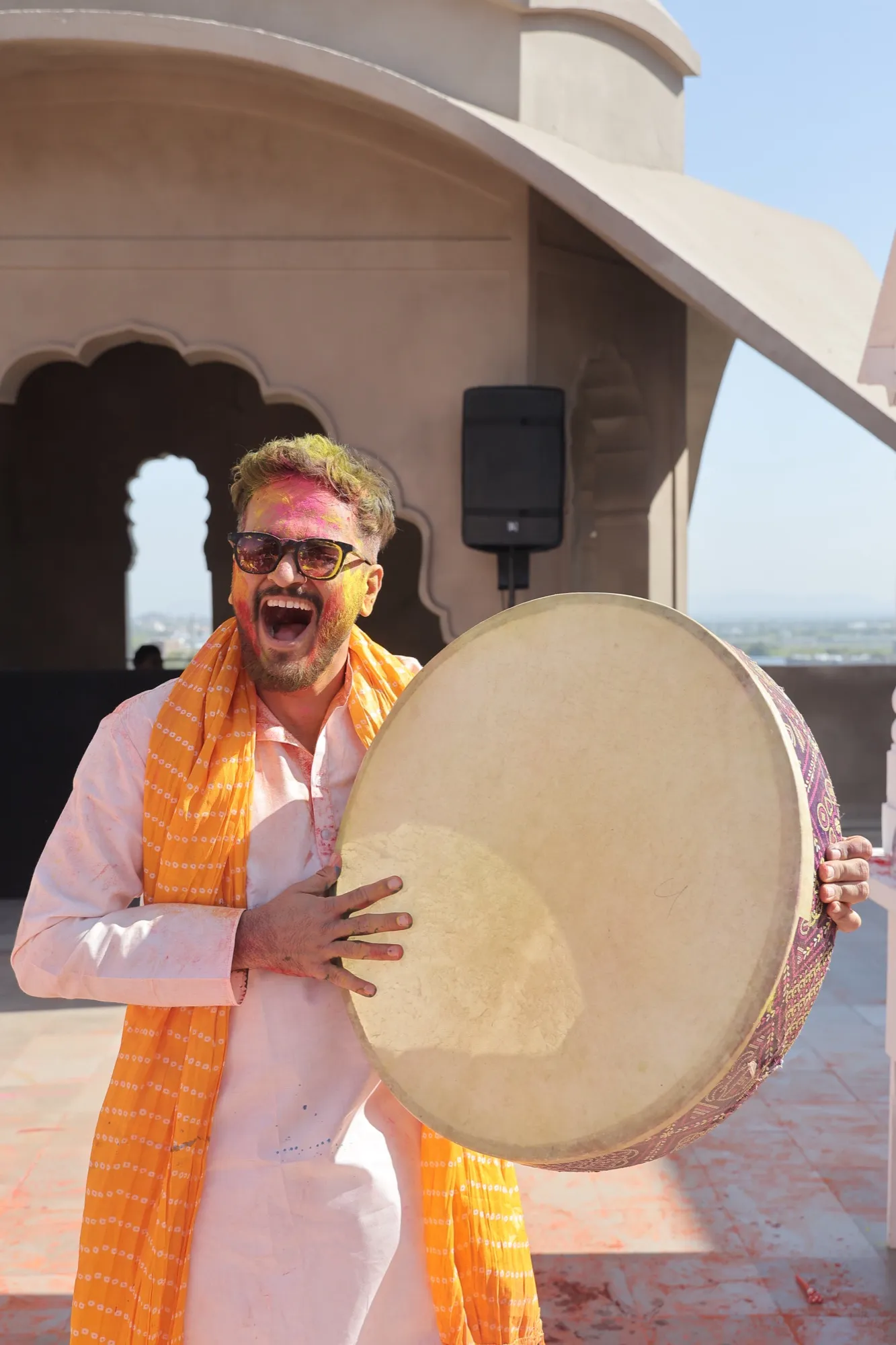 A guest in an orange dupatta laughing as he plays a large frame drum
