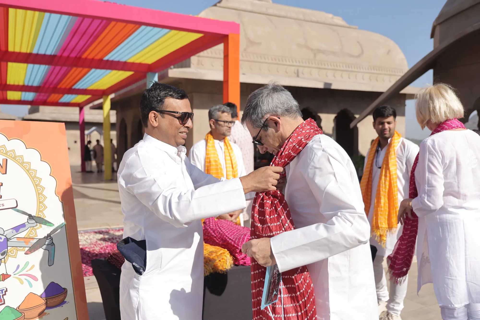 A host tying a red bandhani dupatta around a guest’s neck on the rooftop