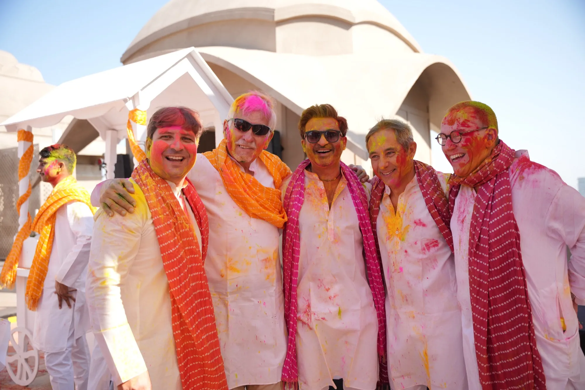 Five guests arm in arm in white kurtas and orange dupattas, all covered in colour
