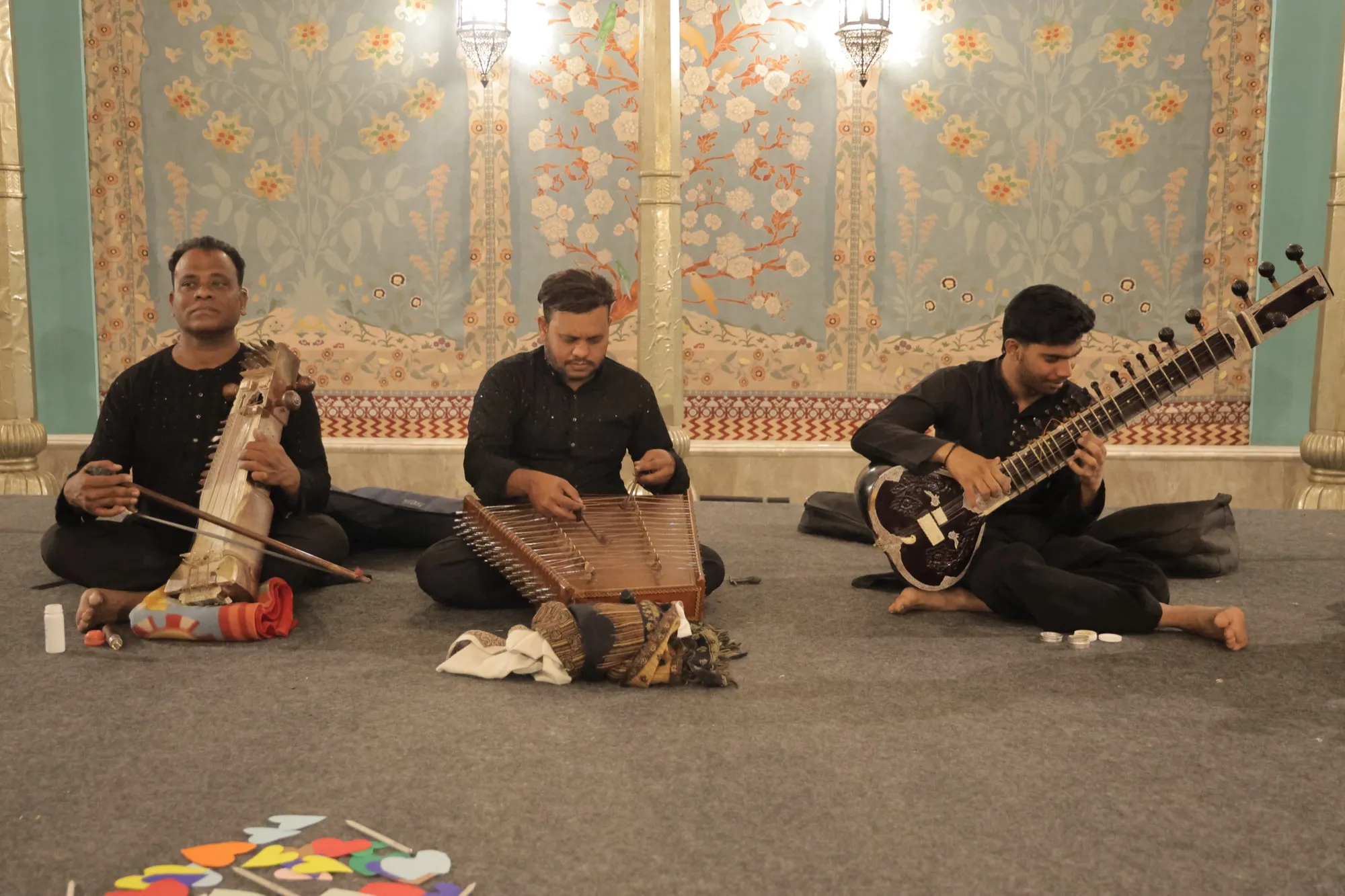 Three musicians seated on the floor playing sarangi, santoor and sitar