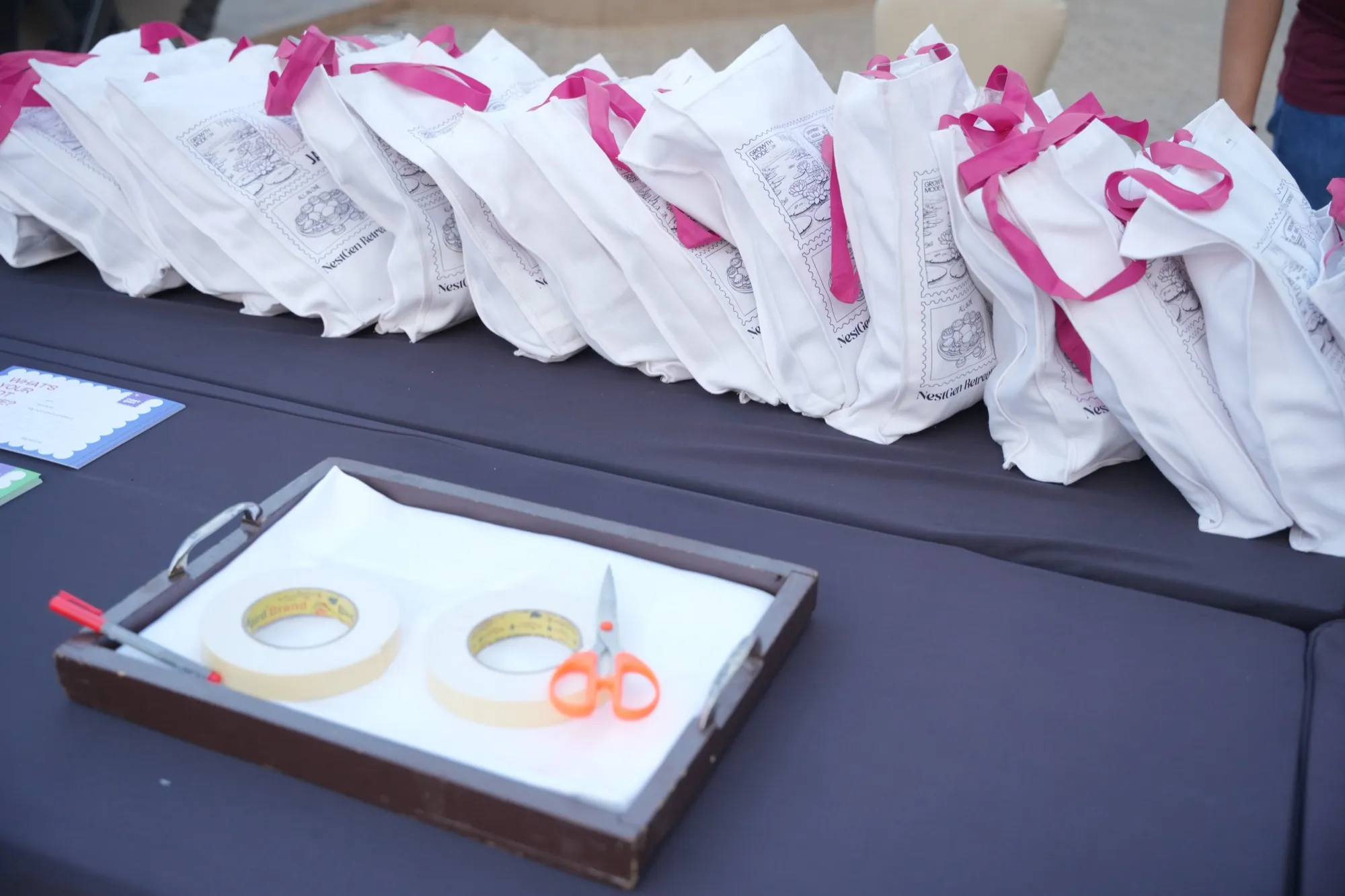 White NestGen tote bags piled on a table, a tray of tape and scissors in front